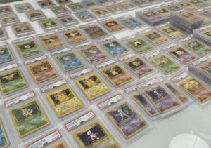 De nombreuses cartes Pokémon sous boîtiers PSA, alignées sur une table avec plusieurs piles de cartes gradées.