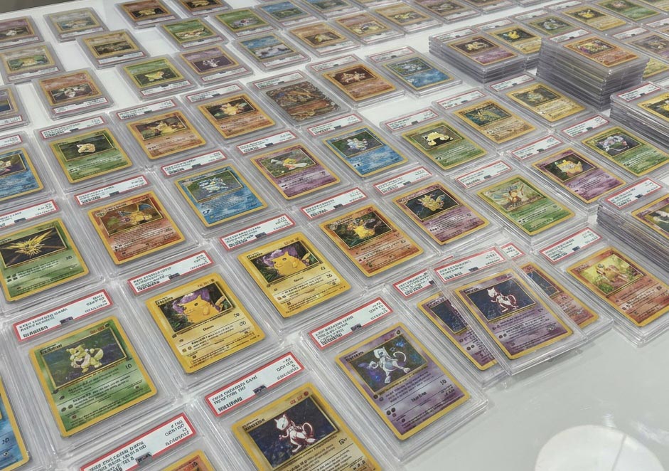 De nombreuses cartes Pokémon sous boîtiers PSA, alignées sur une table avec plusieurs piles de cartes gradées.